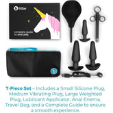 The B-Vibe Anal Training Set of 3 Butt Plugs - Black includes three black silicone plugs in various sizes, an enema bulb, lube applicator, storage pouch, and a unicorn-themed guidebook by B-Vibe, all shown on a white background.